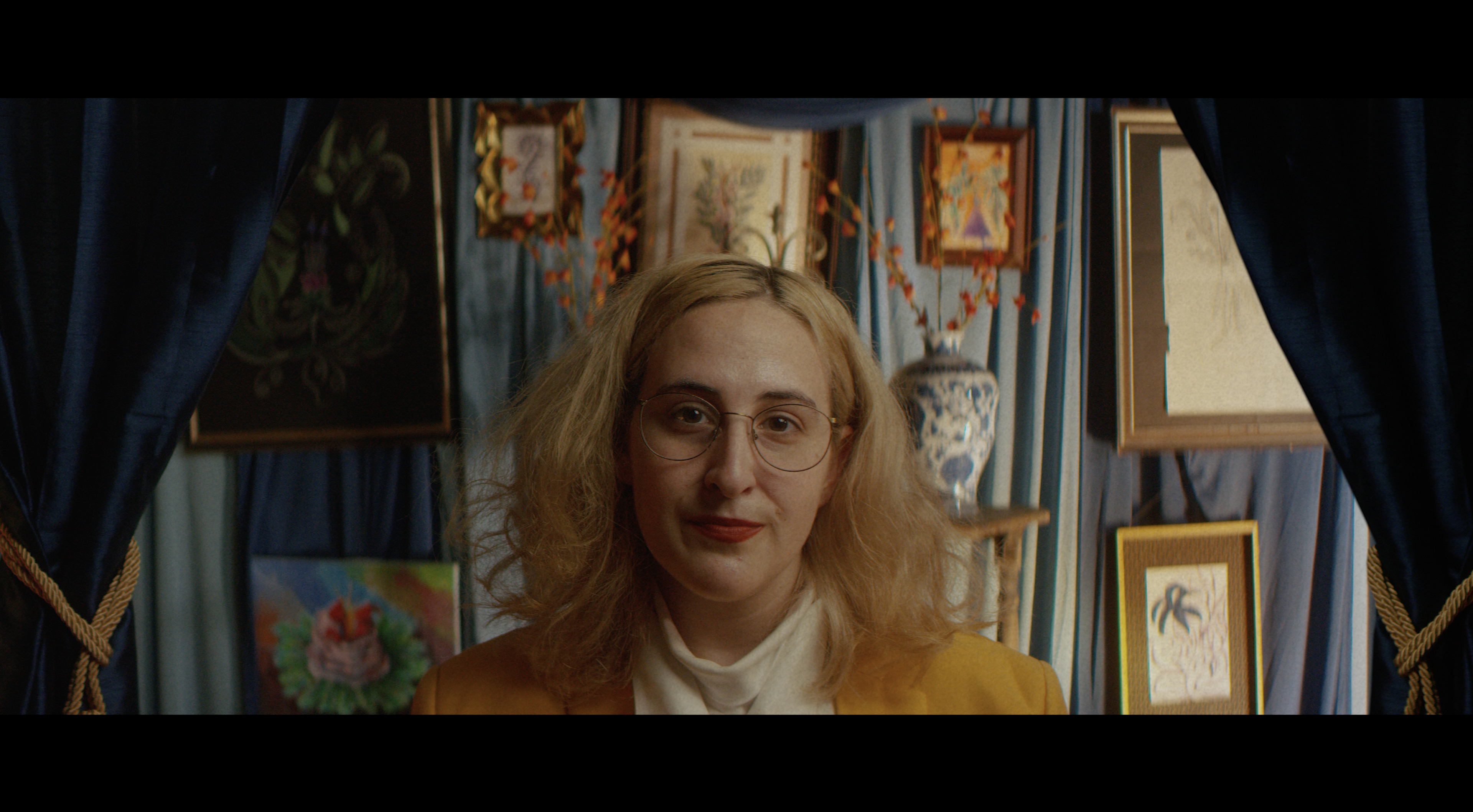 Image Description: A still image from a short film. A person in a yellow suit jacket looks directly into the camera framed by two blue curtains. They stand in front of various artworks.